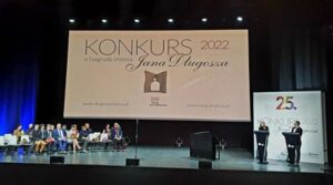 Zdjęcie sceny na której siedzą ludzie. Na środku widoczny jest duży plakat z napisem Konkurs 2022 o Nagrodę imienia Jana Długosza