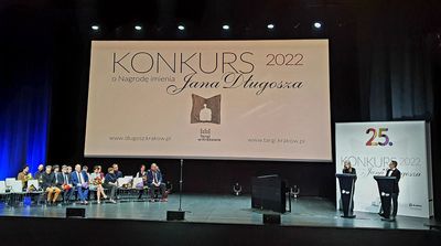 Zdjęcie sceny na której siedzą ludzie. Na środku widoczny jest duży plakat z napisem Konkurs 2022 o Nagrodę imienia Jana Długosza