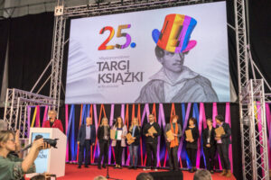 Grupa ludzi stojąca na scenie. Nad nimi duży plakat 25. Targów Książki